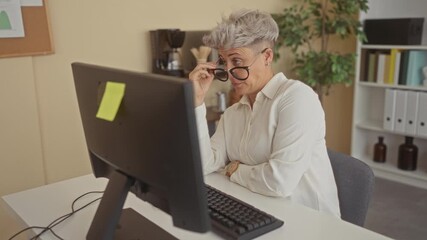 Woman with short hair and glasses sits at office desk by computer monitor and keyboard pressing hand on temple for headache beside coffee maker and potted plant on shelf; fatigue. - Powered by Adobe