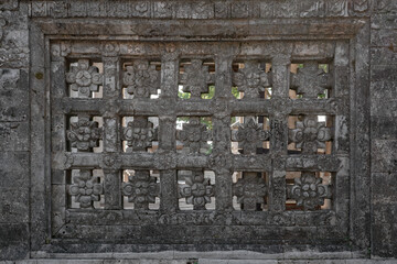 A weathered gray stone wall with intricate traditional Balinese floral carving. An architectural detail of a ventilation grille showing ancient texture.
