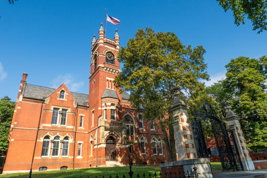 Northampton, MA - US - Aug 24, 2025 A grand brick College Hall at Smith College, with a prominent clock tower and American flag, set against a clear blue sky amidst lush green trees.