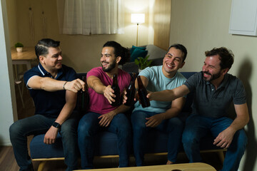 Latin friends toasting with beer bottles watching sports at home