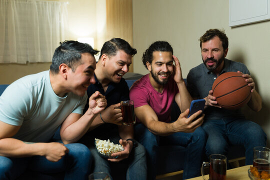 Surprised male friends watching basketball game on smartphone at home