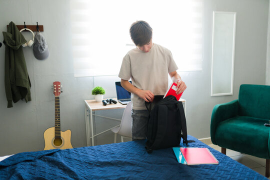 Teenager boy putting notebooks into backpack in bedroom getting ready for school