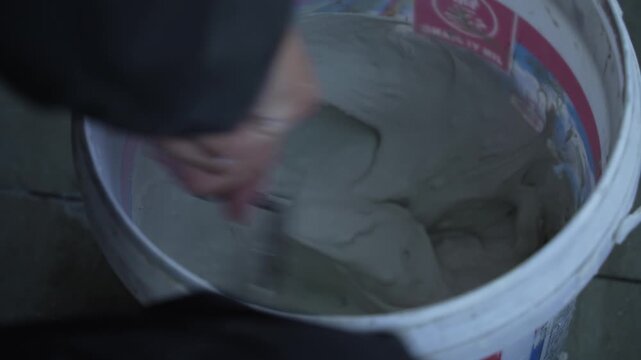 Worker manually stirring white putty paste inside a paint bucket during construction work