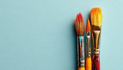 Three paintbrushes with vibrant, multicolored bristles and wooden handles stand against a pale blue background, displaying paint residue