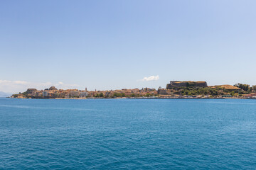 A broad, scenic view of the colorful coastline of Corfu Town, showing a row of historic buildings and the Old Fortress, as seen from the vast, deep blue Ionian Sea on a bright, sunny day.
