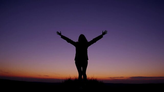 The silhouette of youthful female figure with upper limbs elevated towards the heavens against the sky following solar descent.