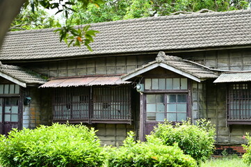 Historic Japanese wooden building with dark tile roof and timber construction surrounded by dense green shrubs, showcasing traditional residential architecture.