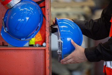 Close-up of hands holding blue safety helmet near storage shelf in an industrial workplace. Concept of PPE usage, workplace safety compliance and personal responsibility in factory environment.