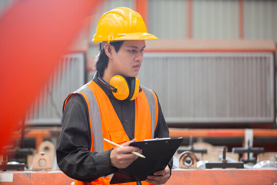 Young Asian factory worker wearing safety vest, helmet, and earmuffs holding clipboard checking for inspection in industrial environment. Concept of safety, engineering and manufacturing.