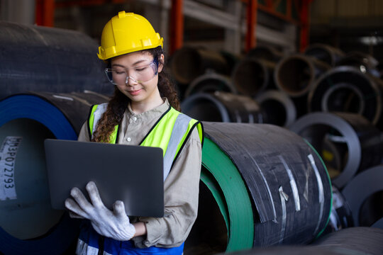Female engineer inspects data on laptop inside metal sheet factory warehouse, focusing on production monitoring, inventory tracking and industrial workflow efficiency.