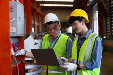 Two industrial engineers wearing safety helmets and reflective vests work together in factory using laptop, focusing on electrical system inspection, data analysis, and equipment monitoring.