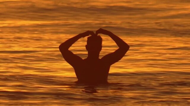 Slow motion close up video of a lone male surfer sits on his surfboard and shows heart shape symbol, silhouetted by the sunset in Indian ocean