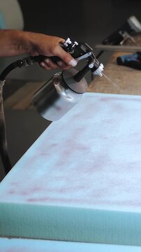 Vertical video. Upholstered furniture factory. Worker applies glue to foam rubber using a spray gun. Gluing foam rubber. The process of assembling and gluing upholstered furniture. Close-up.