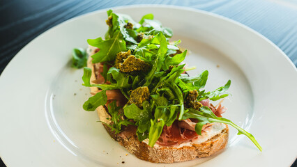 Beautifully served toast with fresh mozzarella, authentic Italian prosciutto, vibrant arugula and green pesto sauce. Perfect for food blogs and menus.