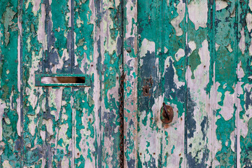 Close-up of a weathered, green wooden door with heavily peeling paint. The rustic texture reveals layers of color, a mail slot, and an old keyhole, creating a perfect vintage or grunge background.

