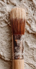 Close-up of a paintbrush with worn, light brown wooden handle and natural bristles resting on a textured beige surface