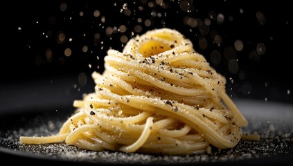 A dark plate holds a high pile of spaghetti, generously seasoned with black pepper and a light dusting of what appears to be salt or cheese, with peppercorns falling onto it