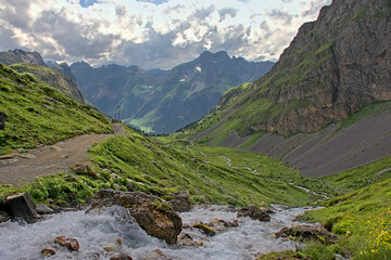Rocky landscape with mountain stream and hiking trail through a green field in La Vanoise national park, France. 