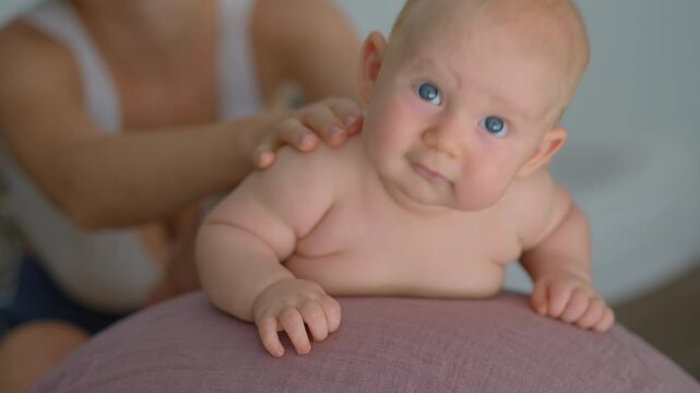 Mother gives a gentle back massage to her 4-month-old baby on a fitball at home. Concept of baby care, bonding, early development, and parent-child interaction.