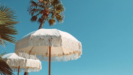 Low-angle view of two elegant white beach umbrellas with fringed edges, under a vibrant blue sky and palm trees