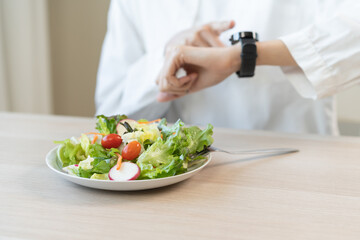 Intermittent fasting, diet healthy asian young woman dietary watching wrist watch, temptation hungry of brunch food in morning on table at home, waiting time to eat salad but not yet time first meal.