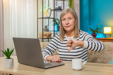 Mature woman reading negative comment on laptop, shaking head and giving thumbs down dislike...