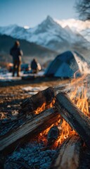 A crackling campfire dominates the foreground, with blurred figures and a tent amidst a snowy mountain landscape at sunset