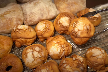 Assorted bakery bread rolls with sesame topping on a cooling rack, with tongs picking one up