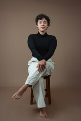 Pretty young woman, no make up on, barefoot, sitting on a stool with crossed legs and hands on knee. Elegant pose for Gen Z attitude, self confidence, female empowerment. Studio portrait, copy space.