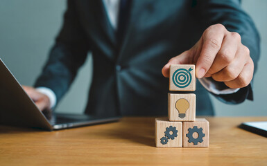 Businessman stacking wooden blocks with target, lightbulb, and gear icons, representing business strategy, goal setting, and innovative solutions.