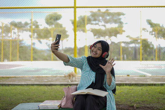 Enthusiastic young woman wearing a hijab and headphones,  happily waving into her smartphone for a selfie or video call, reflecting modern connection, personal joy, and casual student interaction
