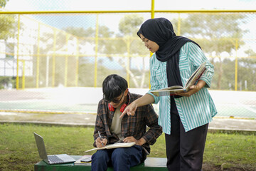 Two students studying together outdoors, one pointing at a notebook while the other writes.  Collaboration, learning, education, teamwork, and friendship are key themes