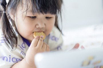 Cute little Asian girl sitting on the bed eating cookies and looking at the tablet,Asian cute...
