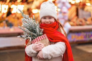 Child enjoys holiday festivities at a market while holding a miniature Christmas tree wrapped in red scarf and warm clothing