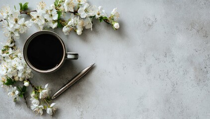 Flatlay of dark coffee in a mug, silver pen, and blooming white cherry blossoms arranged on a light gray textured surface, leaving ample space