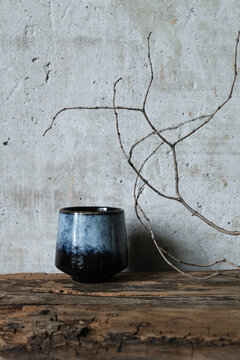 Minimalist still life with a dark ceramic vase and dry branch on rustic wood against a textured concrete wall. Wabi-sabi style composition