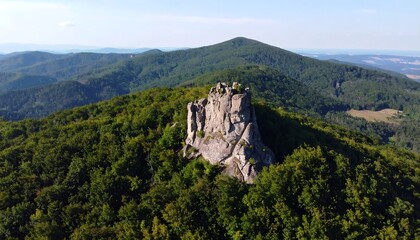 A towering rock formation stands prominently amidst a dense forest-covered mountain range.