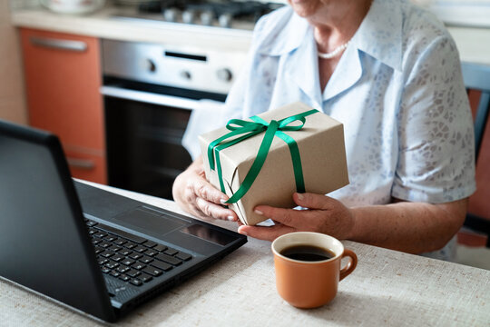 Elderly woman holding gift box, using laptop at home kitchen for video call, online connection. Senior virtual celebration alone, presents sharing, holiday parcel. Technology lifestyle in retirement - Powered by Adobe
