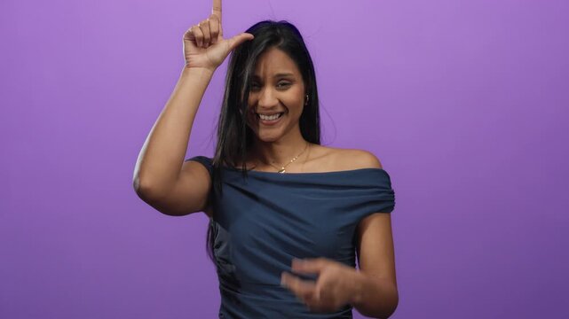 Young hispanic woman makes loser gesture against an isolated purple background, smiling playfully at the camera, conveying a relaxed and humorous atmosphere.