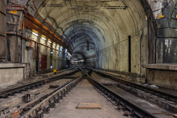 A powerful perspective shot of intersecting train tracks inside a grimy, illuminated tunnel. The converging lines and arched ceiling create a sense of mystery and urban exploration.