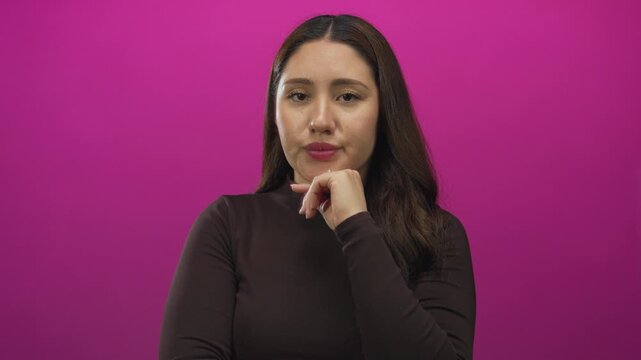 Woman holds hand on chin with direct gaze at camera for thinking in studio against pink wall; introspection.