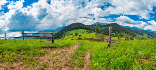 Rustic wooden farm gate and dirt path to a Carpathian village, panoramic summer landscape with green meadows and forested mountains, Ukraine