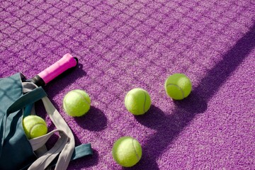 A teal sports bag with a pink-gripped racket and several yellow balls scattered across a purple padel court