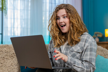 Young woman at home sitting with laptop suddenly raising eyebrows surprised by unexpected positive...
