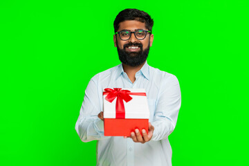 Indian man extends hands holding a gift box with smile, inviting someone to take it as a generous surprise. Arabian guy isolated on green chroma key background showing kindness happy giving gesture.