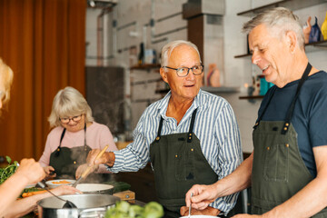 Smiling senior man stirring pot while talking with male friend during cooking class in commercial kitchen