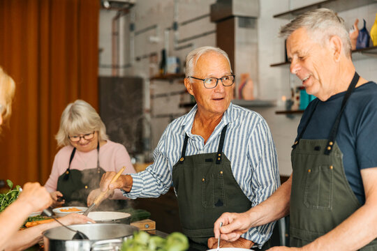 Smiling senior man stirring pot while talking with male friend during cooking class in commercial kitchen