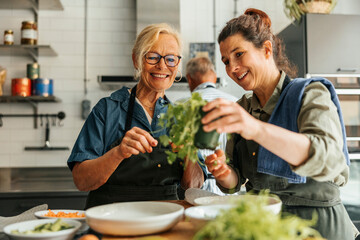 Smiling female chef holding basil plant while talking with senior woman during cooking class in commercial kitchen