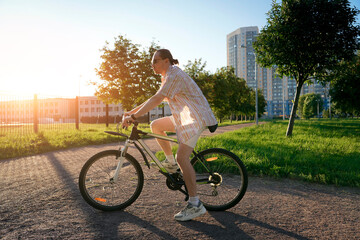 Obraz premium A man with a ponytail is riding a bicycle along a paved path in a park. The sun is setting and creating a golden hour light, casting a flare in the sky. Tall buildings are in the distance