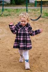 Cute caucasian blond kid in a pink and black plaid coat stands on playground sand. Outdoor toddler play and development. © Serg